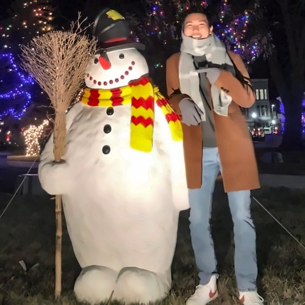 Min next to a giant snowman among holiday lights at night