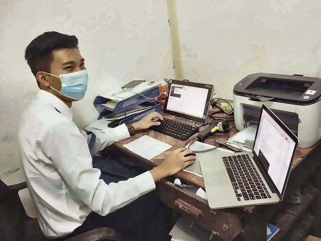 Min working at a desk with two laptops, wearing a face mask